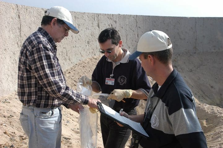 Three men from the IAEA hold test samples in plastic bags. They are standing in a pit filled with sand.