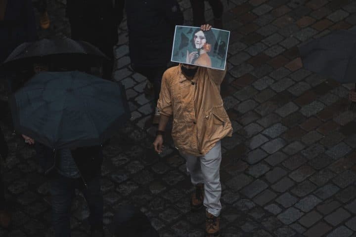 A lone person wearing a brown jacket holds up a picture of Mahsa Amini. People around him wear all black and carry umbrellas.