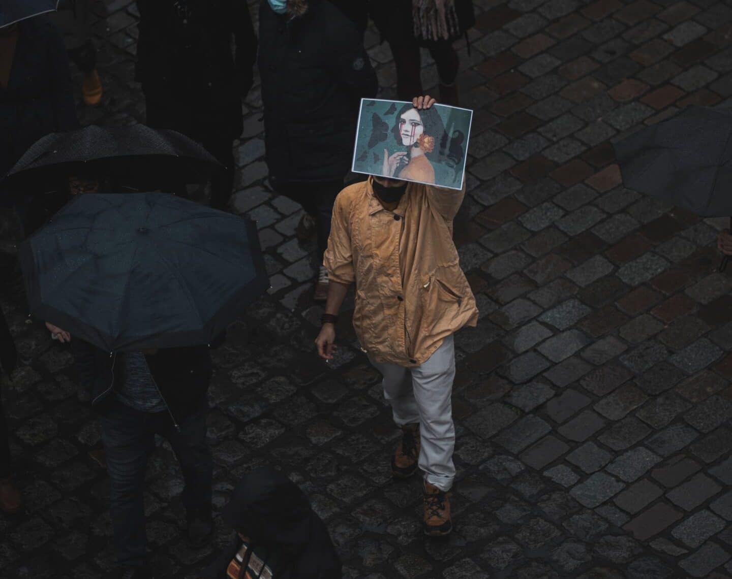 A lone person wearing a brown jacket holds up a picture of Mahsa Amini. People around him wear all black and carry umbrellas.