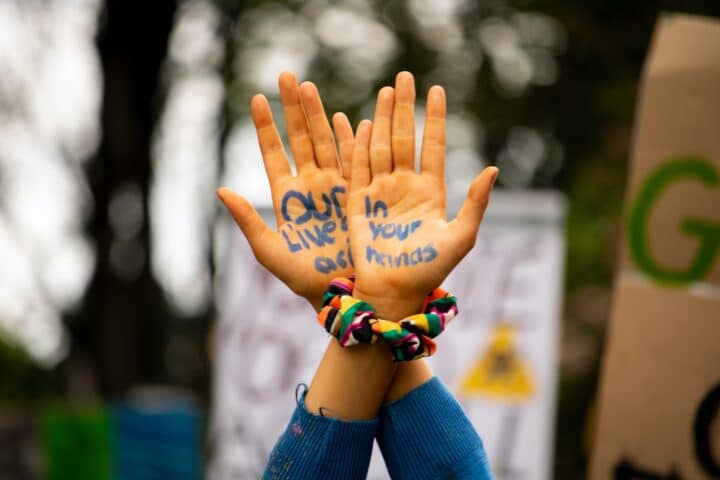 Two hands are crossed and held aloft to the camera. On the palms are the words "Our lives are in your hands". Behind the hands are placards, indicating a protest.
