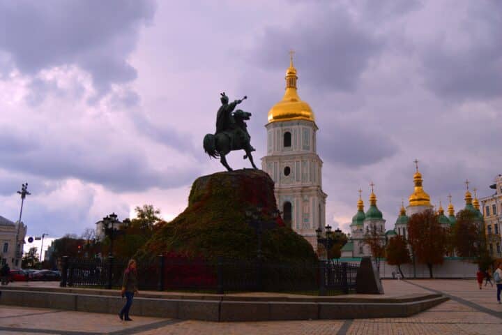 A statue of a man riding a horse in a square in Kiev, Ukraine. Behind the statue is a large monastery with golden domes.