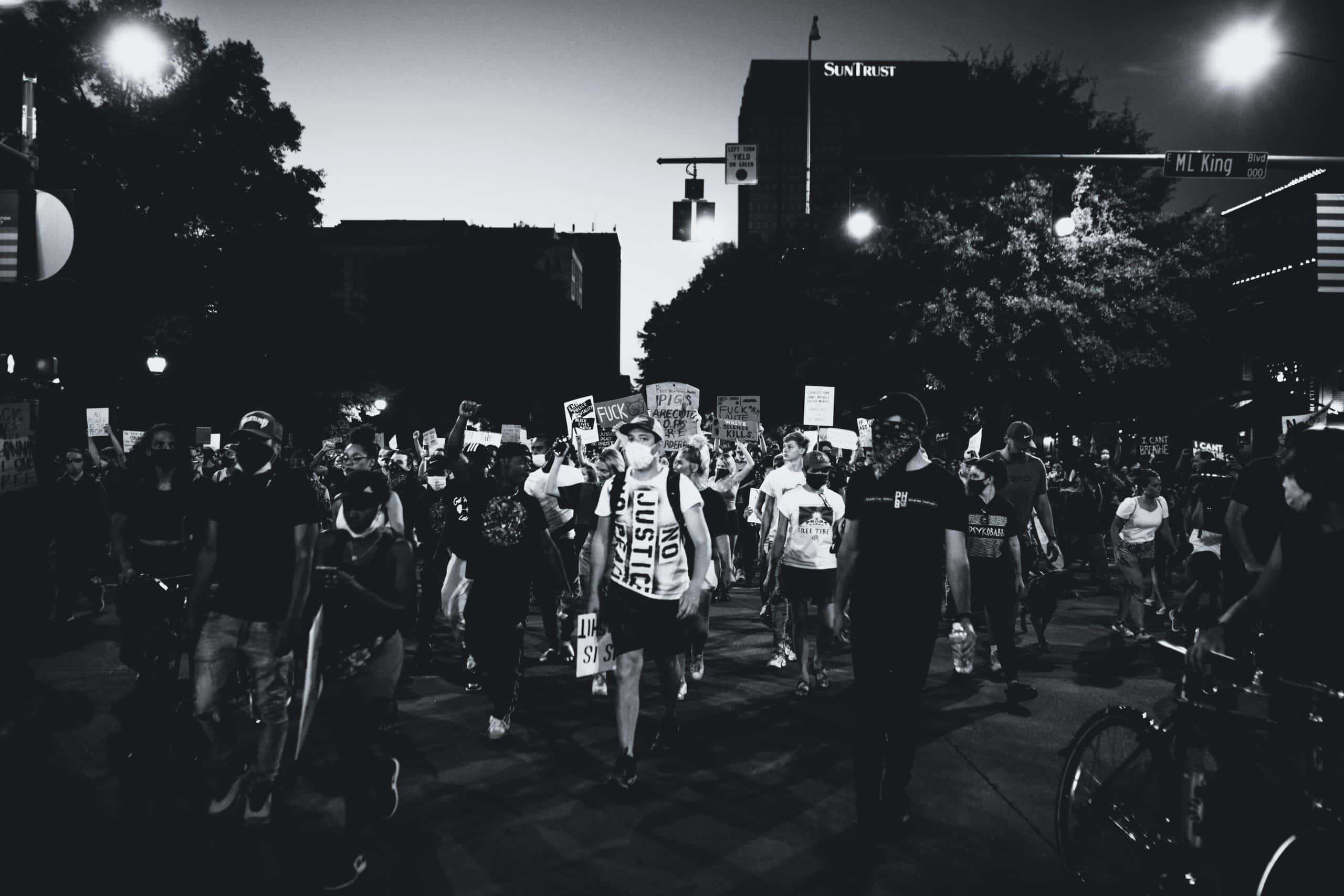 Black and white image of protesters marching down Martin Luther King Boulevard. Many have signs denouncing the police in America; most are wearing face masks.