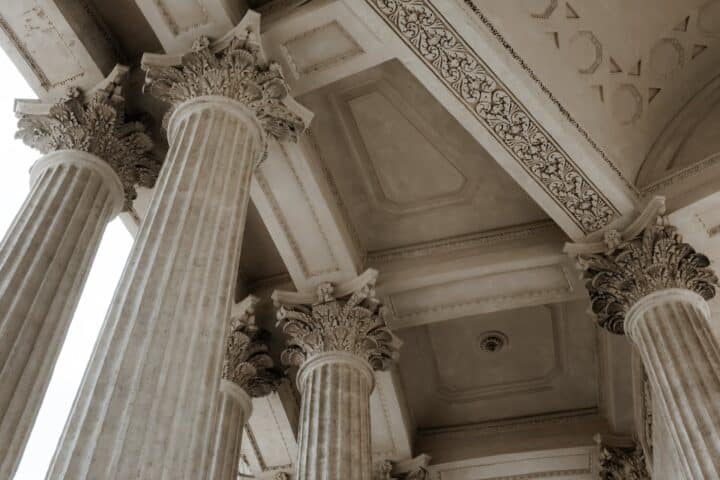 The ceiling of a building is shown, built in a classical fashion which mimics the architecture of the ancient Greeks, with tall columns holding up the roof.