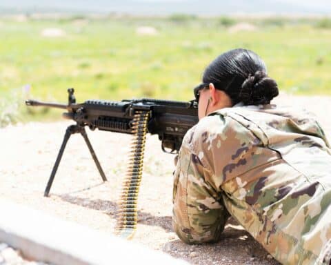 A woman in a military uniform is lying prone, aiming an automatic weapon.