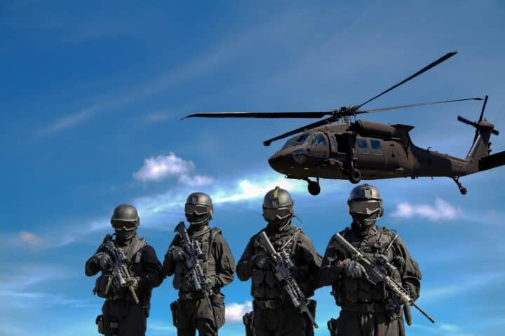 Four masked soldiers holding rifles look at the camera while a helicopter flies closely overhead.