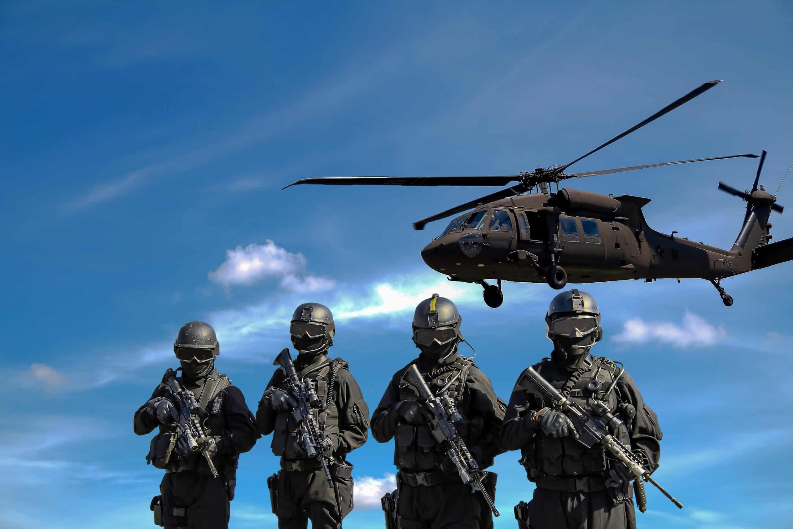 Four masked soldiers holding rifles look at the camera while a helicopter flies closely overhead.