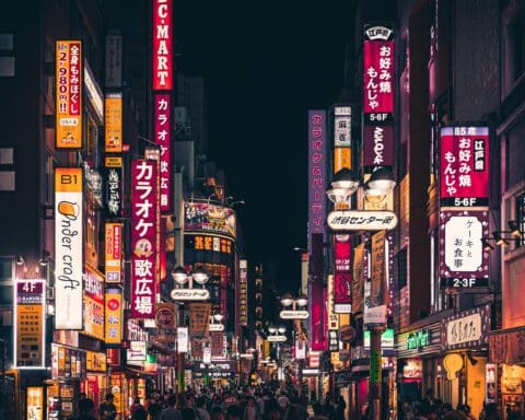 People walking down a busy, narrow street in Tokyo, Japan. Brightly coloured neon signs occupy the walls of the buildings on the street.