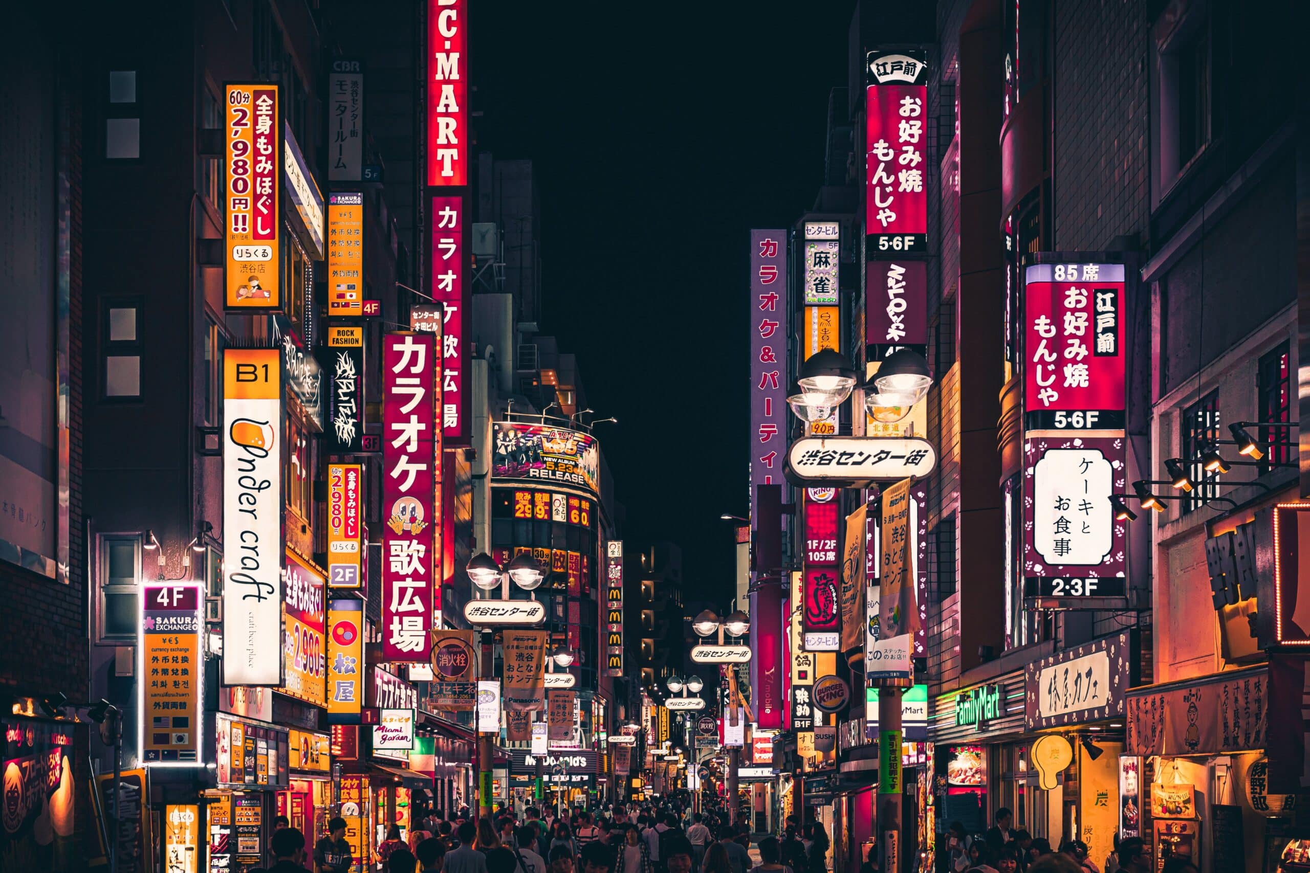 People walking down a busy, narrow street in Tokyo, Japan. Brightly coloured neon signs occupy the walls of the buildings on the street.