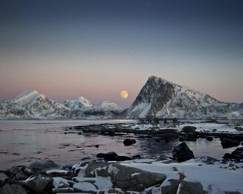 A full moon shines across an arctic landscape, peeking through snow-capped mountains. Beneath them is icy water.