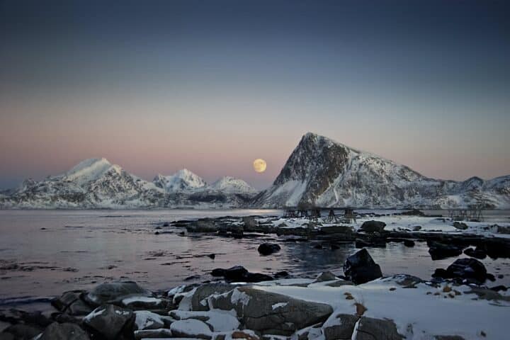 A full moon shines across an arctic landscape, peeking through snow-capped mountains. Beneath them is icy water.