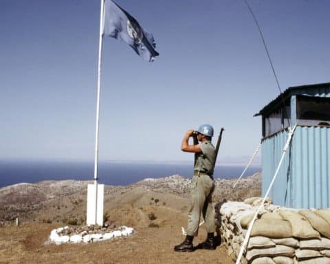 A lone U.N peacekeeping soldier stands atop a mountain, looking down with binoculars on the hills of Cyprus. Surrounding him is a small guard shack and a flagpole bearing the U.N flag.
