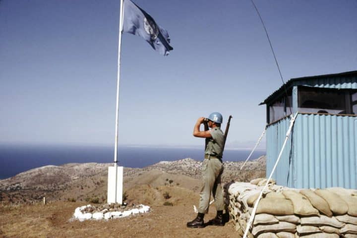 A lone U.N peacekeeping soldier stands atop a mountain, looking down with binoculars on the hills of Cyprus. Surrounding him is a small guard shack and a flagpole bearing the U.N flag.