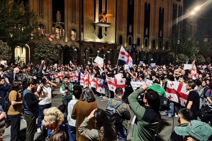 Protests in Tbilisi, Georgia, in April of 2024. A large crowd is marching down a street, holding many Georgian flags. Opposite them stand several reporters and photographers, taking pictures. Behind them is a tall and very grand building.