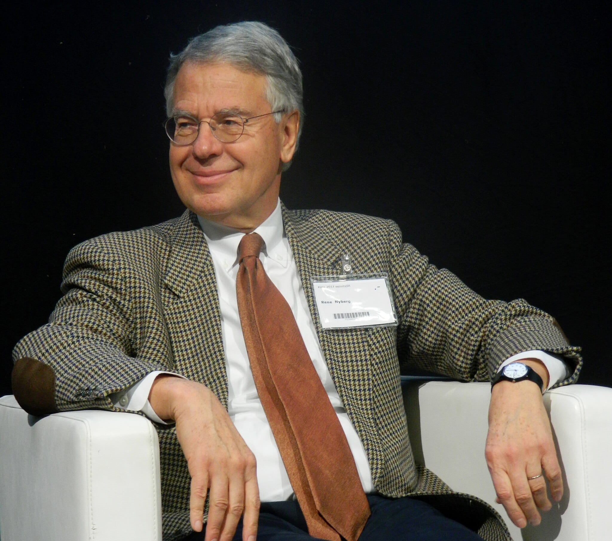 Ambassador René Nyberg, dressed in a suit with an orange tie, sits and smiles in an armchair while speaking at the Helsinki bookfair in 2017.