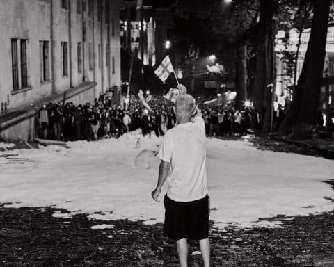 A lone man waving a small Georgian flag on a snow-covered street. In the distance a large crowd is marching towards him.