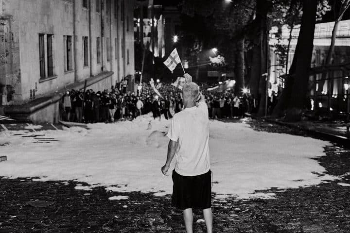 A lone man waving a small Georgian flag on a snow-covered street. In the distance a large crowd is marching towards him.