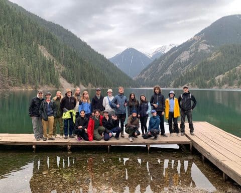 A large group of people, the UF Uppsala Travel Group, stands on a pier, looking at the camera. Behind them is a lake with large mountains towering over them.