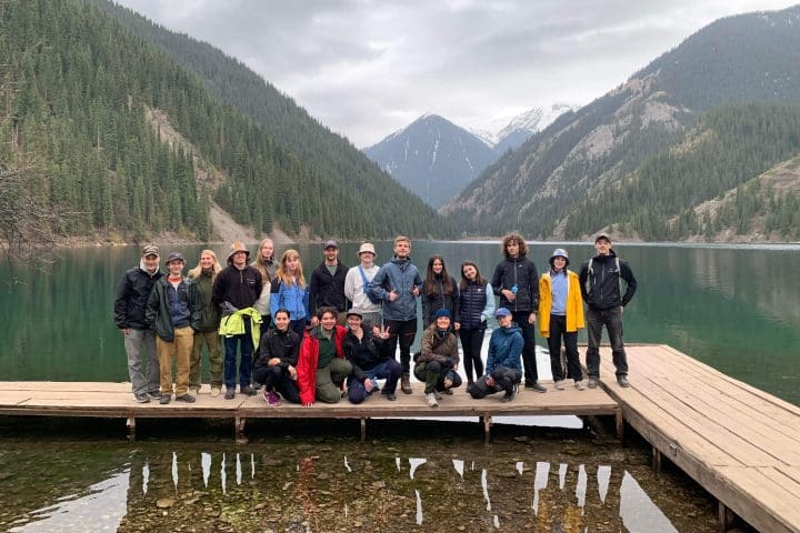 A large group of people, the UF Uppsala Travel Group, stands on a pier, looking at the camera. Behind them is a lake with large mountains towering over them.