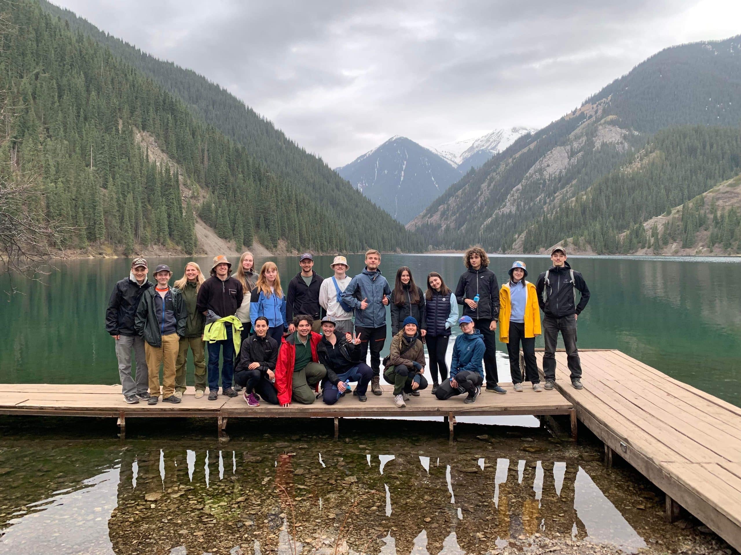 A large group of people, the UF Uppsala Travel Group, stands on a pier, looking at the camera. Behind them is a lake with large mountains towering over them.