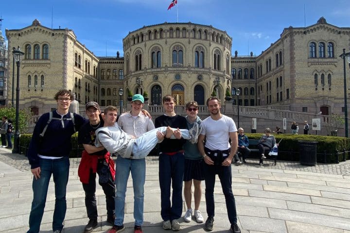 The members of the UF Uppsala Travel Group standing in front of the Oslo Parliament building. One of the members is being held aloft by another, in a comic pose.