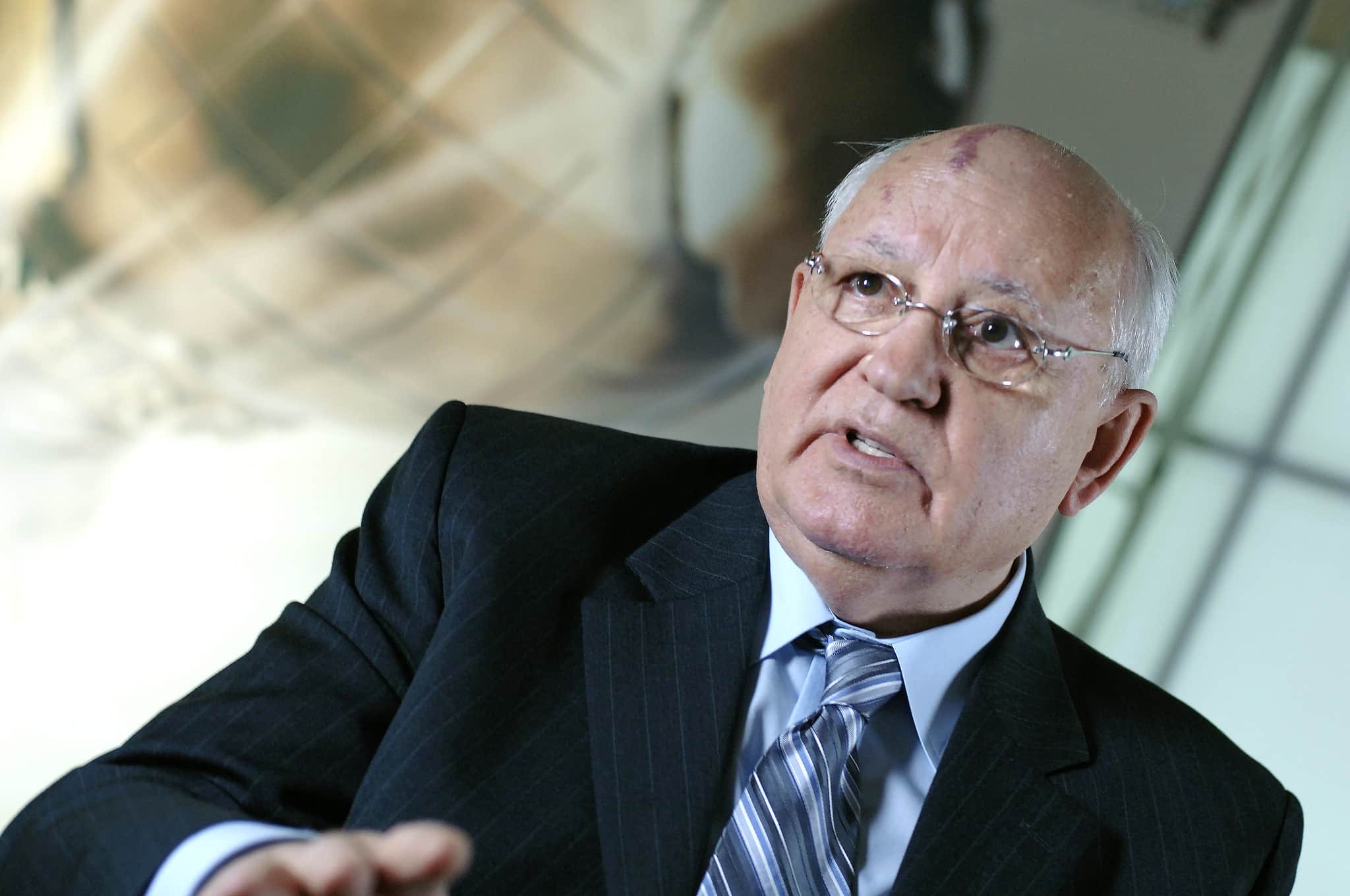 An older Mikhail Gorbachev, dressed in a dark suit with glasses on, is speaking.