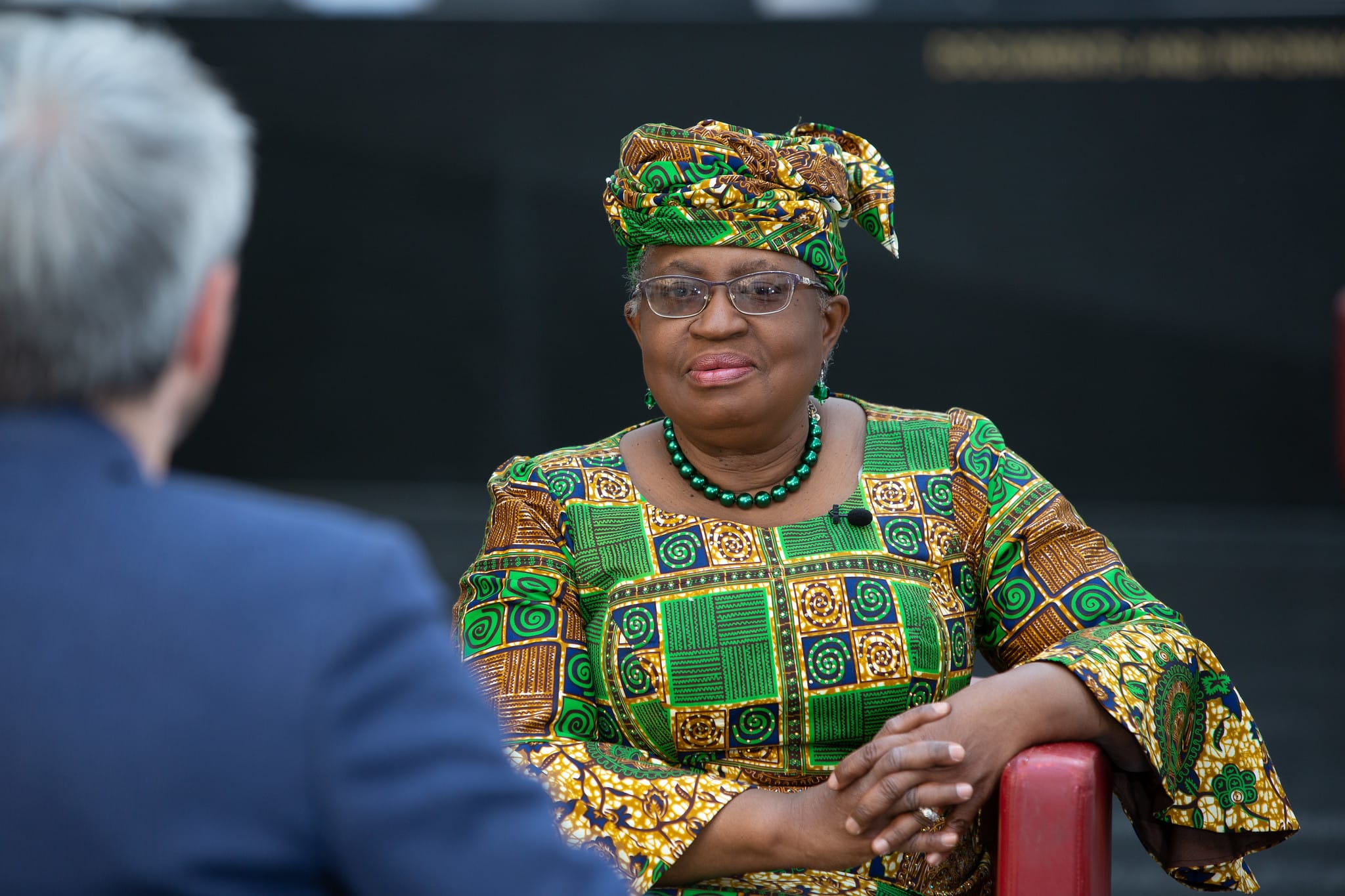 Ngozi Okonjo-Iweala, dressed in a traditional African dress, sits in an armchair before a man in a suit. They appear to be conversing, though Ngozi is only listening.