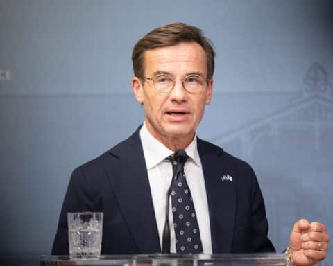 Swedish Prime Minister Ulf Kristersson, dressed in a dark suit with a dotted tie, speaks behind a podium. The wall behind him is blue and on the podium stands a small microphone and a glass of water. He is speaking and gesturing with his hand.