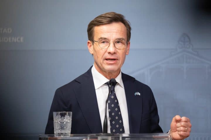Swedish Prime Minister Ulf Kristersson, dressed in a dark suit with a dotted tie, speaks behind a podium. The wall behind him is blue and on the podium stands a small microphone and a glass of water. He is speaking and gesturing with his hand.