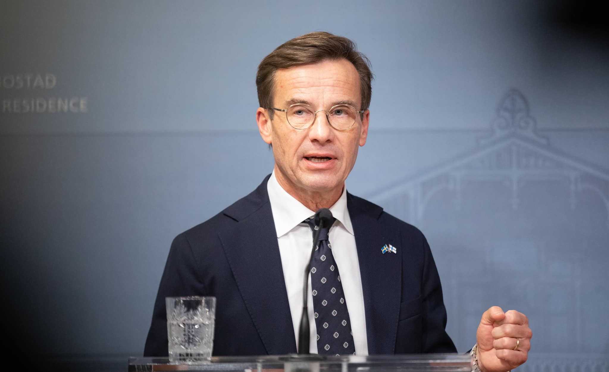Swedish Prime Minister Ulf Kristersson, dressed in a dark suit with a dotted tie, speaks behind a podium. The wall behind him is blue and on the podium stands a small microphone and a glass of water. He is speaking and gesturing with his hand.