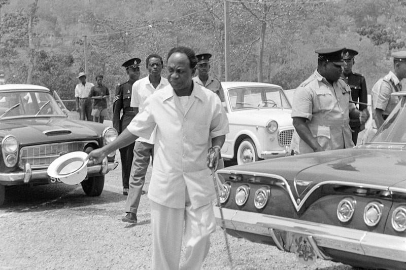 President Kwame Nkrumah in white western clothes is visiting waving with a tropical hat and a cane. Five smiling policemen and three passenger cars. The tailgate of a 1961 Chevrolet Impala presidential car with six red and white lights and a Ghanian star.