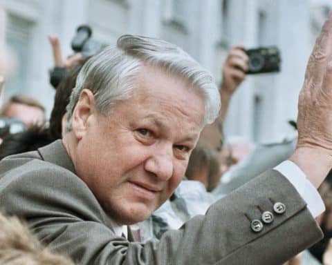 President of Russia Boris Yeltsin waves at reporters, who hold their cameras aloft behind him.