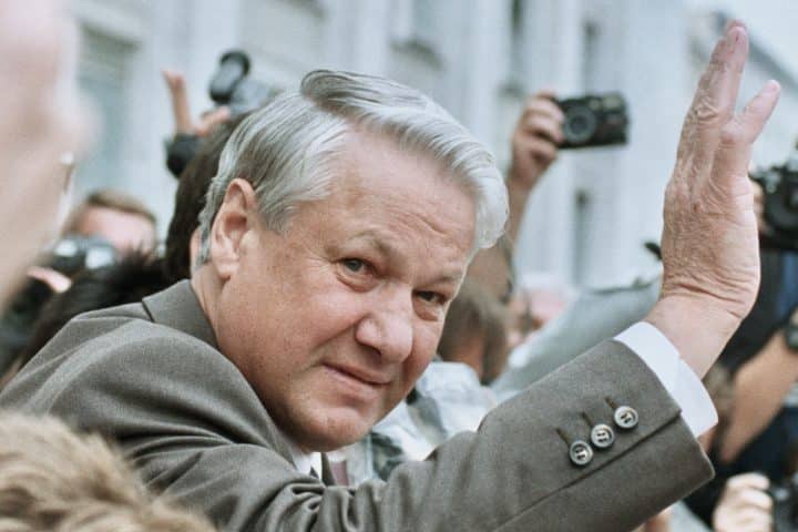 President of Russia Boris Yeltsin waves at reporters, who hold their cameras aloft behind him.