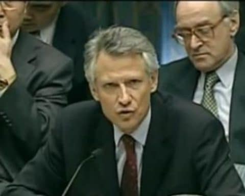 Then French Minister of Foreign Affairs Dominique de Villepin gives his famous speech at the U.N, seated in a dark suit with a red tie. Foreign diplomats sit behind him in several rows as he speaks into a microphone.