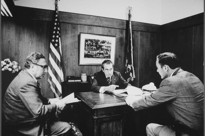 Secretary of State Henry Kissinger with President Nixon and General Haig, discussing Vietnam. Nixon sits behind a desk while the others face him. Behind them are two American flags and a hanged photograph of horses. Black and white photograph.