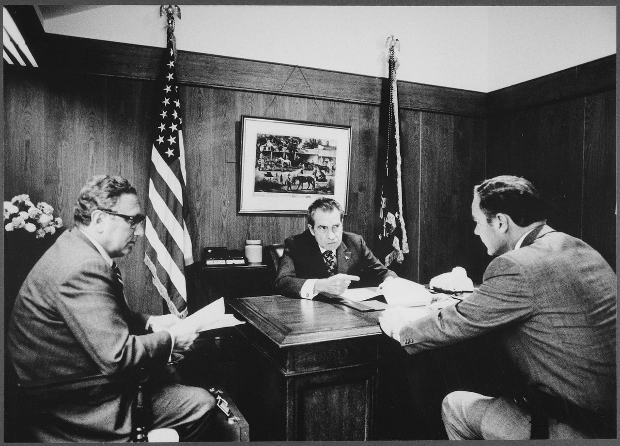 Secretary of State Henry Kissinger with President Nixon and General Haig, discussing Vietnam. Nixon sits behind a desk while the others face him. Behind them are two American flags and a hanged photograph of horses. Black and white photograph.