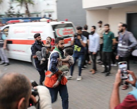 A man rushes a wounded and soot-covered child to hospital, with another man carrying another child directly behind him. Around them stands a crowd, with an ambulance in the background.