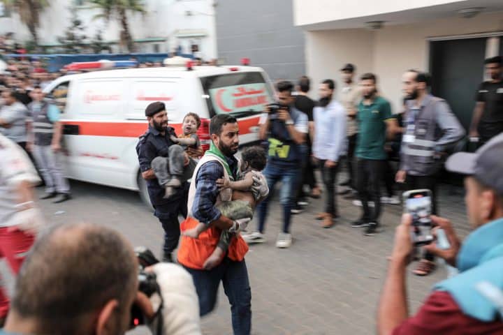 A man rushes a wounded and soot-covered child to hospital, with another man carrying another child directly behind him. Around them stands a crowd, with an ambulance in the background.