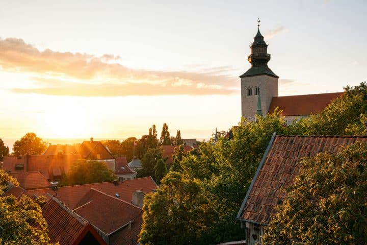 Solen sätter sig över Visby, Gotland. En kyrka är tydligt synlig och taken av flera hus, med träd kring sig. Här finns Almedalsveckan.