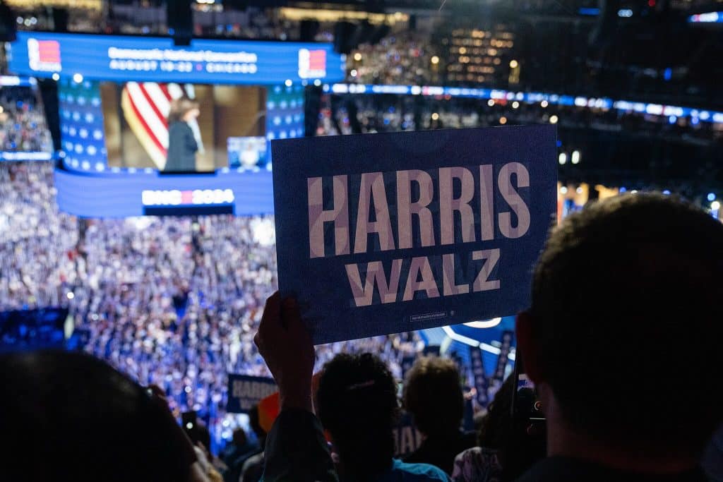 Kamala Harris can be seen taking the stage at the 2024 Democratic National Convention on a large TV-screen. A massive crowd is cheering her on. The stage itself is blocked from view by someone holding a blue "Harris Walz" sign close to the camera.