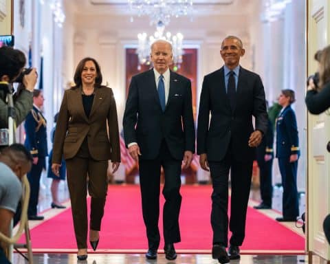 From left to right: Vice President Kamala Harris, current President Joe Biden, and former President Barack Obama. The three are walking towards the camera taking the photo, out of an elegant room into a corridor packed with journalists and cameras. Their stride is confident, and they are all smiling.