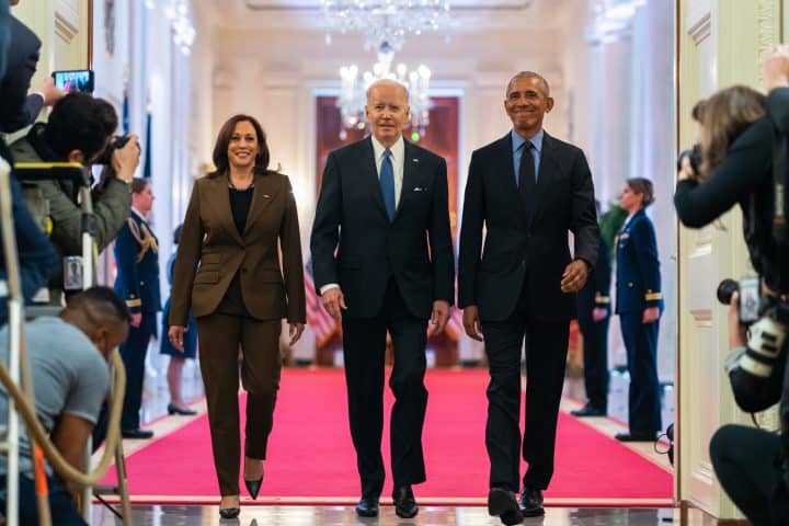 From left to right: Vice President Kamala Harris, current President Joe Biden, and former President Barack Obama. The three are walking towards the camera taking the photo, out of an elegant room into a corridor packed with journalists and cameras. Their stride is confident, and they are all smiling.