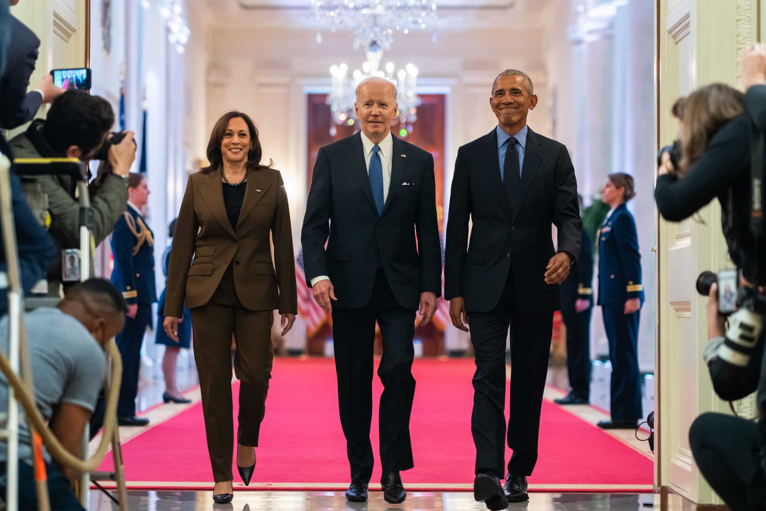 From left to right: Vice President Kamala Harris, current President Joe Biden, and former President Barack Obama. The three are walking towards the camera taking the photo, out of an elegant room into a corridor packed with journalists and cameras. Their stride is confident, and they are all smiling.