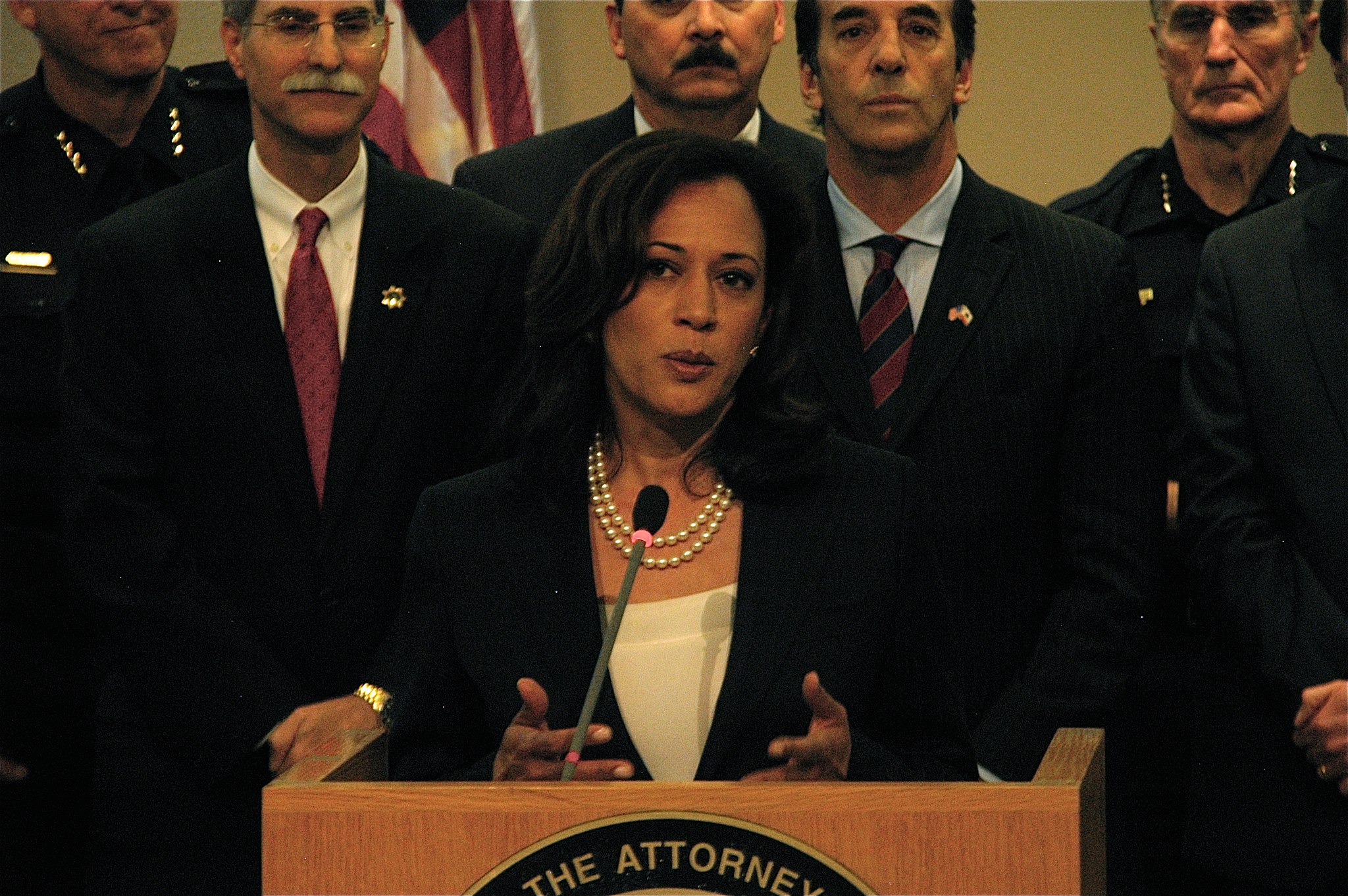Kamala Harris is speaking from a podium, during her time as Attorney General in California. She has a serious face and is gesturing with her hands. Behind her stand police officers and other officials.
