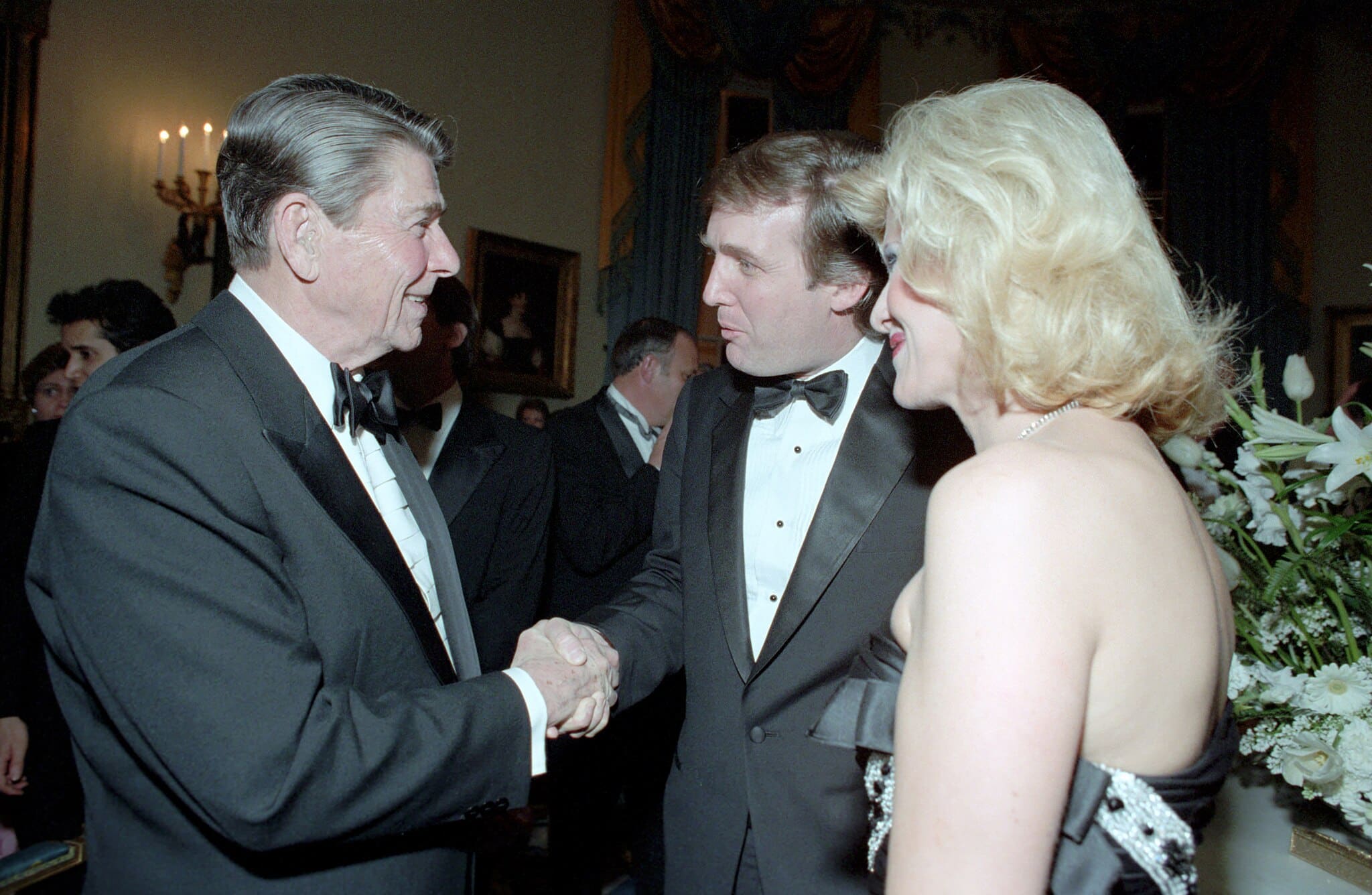 Donald Trump shaking hands with President Ronald Reagan during a State Dinner in 1985 at the White House. Both are wearing black suits, white shirts and black ties. Donald Trump's then wife Ivana looks on in a black dress. Everyone is smiling.