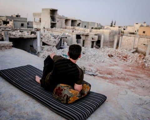 A young man is lounging on a carpet, with a pillow under his arm, in a bombed out village, showing that peace can be found even amidst destruction.