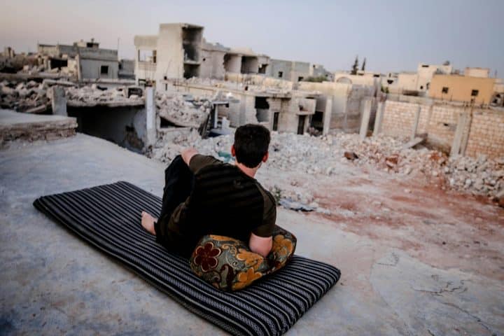 A young man is lounging on a carpet, with a pillow under his arm, in a bombed out village, showing that peace can be found even amidst destruction.
