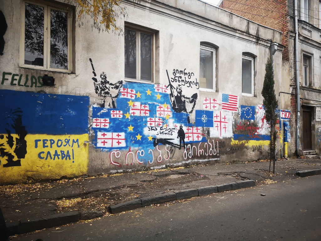 The wall of a building covered in graffiti. There are Georgian, Ukrainian, EU and NATO flags, along with soldiers and various text both in Georgian and English.