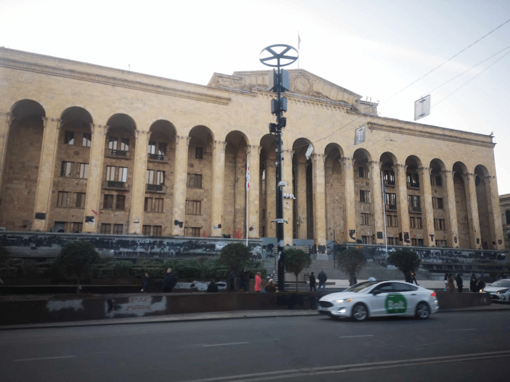 The parliament building in Tbilisi and the street in front of it. The concrete foundation and bottom of the pillars are covered in black paint, masking graffiti underneath.