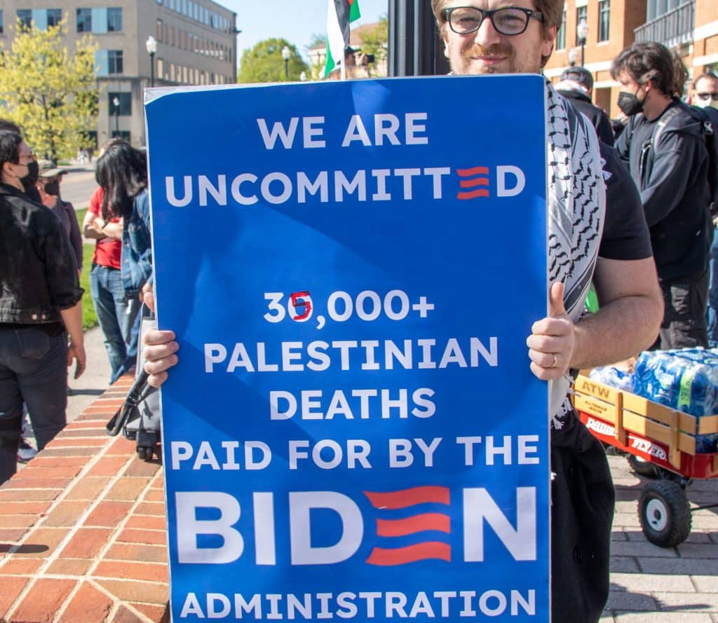 A man at a pro-Palestinian protest holds a sign which reads: "WE ARE UNCOMMITTED. 35,000+ PALESTINIAN DEATHS PAID FOR BY THE BIDEN ADMINISTRATION". The sign is blue and is made to look similar to an official Biden-Harris campaign sign.
