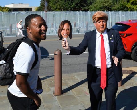 An imitator dressed as Donald Trump waves a picture of Kamala Harris on a stick, while a man standing closely to the imitator laughs.